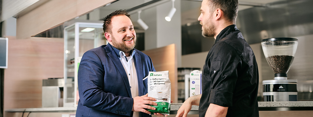 Zwei Personen stehen in einem Bistro-Bereich mit der Aufschrift „barista & bar“ und halten Verpackungen mit Kaffeeprodukten, während sie sich unterhalten.