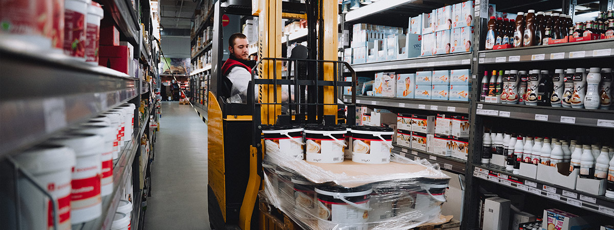 Eine Person in Selgros-Arbeitskleidung fährt mit einem Gabelstapler durch die Lagergänge, transportiert palettierte Waren, während Regale mit Produkten zu sehen sind.