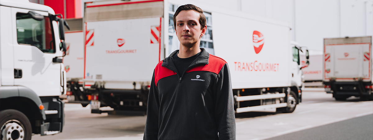 Eine Person steht in Arbeitskleidung auf dem Betriebsgelände von Transgourmet Deutschland, umgeben von Lieferfahrzeugen.