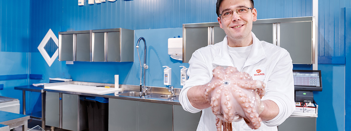 Eine Person in Arbeitskleidung steht hinter der ‚Frischfisch‘-Theke und präsentiert einen Tintenfisch.