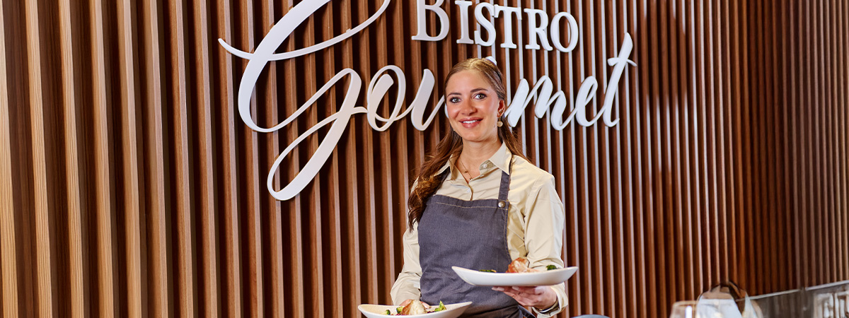 Eine Person im Bistro Gourmet trägt eine Schürze und hält zwei Teller mit Speisen, während im Hintergrund an der Holzvertäfelung die dekorative Schrift "Bistro Gourmet" zu sehen ist.