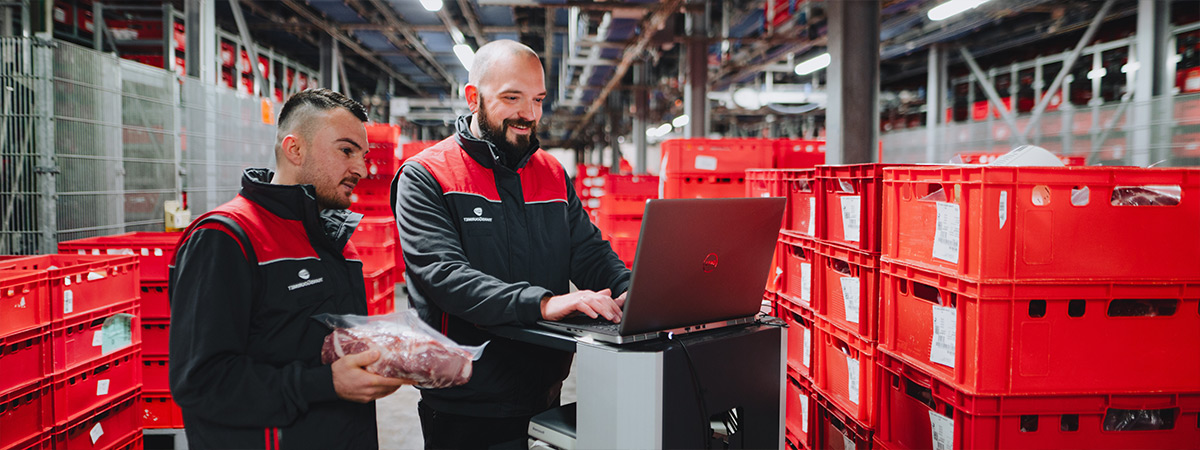 Zwei Personen in Arbeitskleidung arbeiten in einer Lagerhalle, eine hält ein verpacktes Stück Fleisch, während die andere an einem Laptop tätig ist.