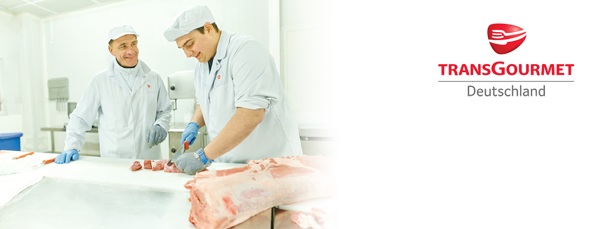 Zwei Personen stehen in weißer Arbeitskleidung in einer industriellen Umgebung. Eine Person schneidet ein Stück Fleisch.