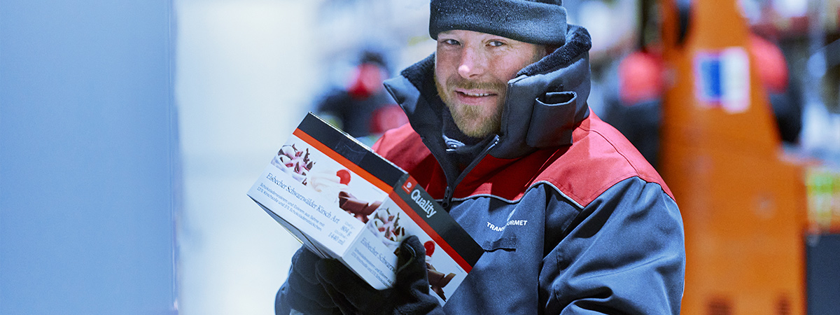 Eine Person steht in warmer Arbeitskleidung in einem Tiefkühllager und hält ein abgepacktes Lebensmittel in den Händen.