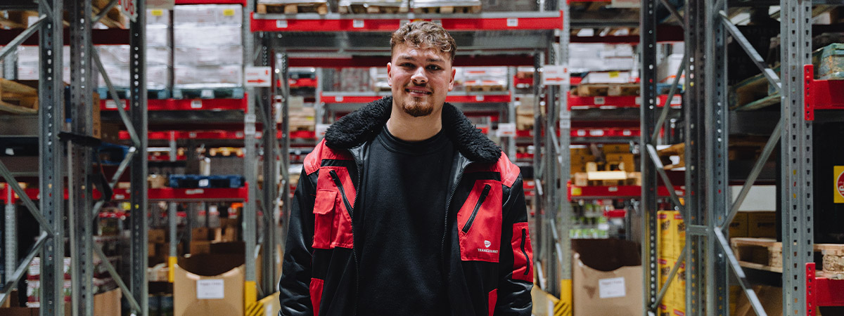 Eine Person steht in einem Lagerbereich mit Regalen voller Waren, gekleidet in einer Arbeitsjacke.