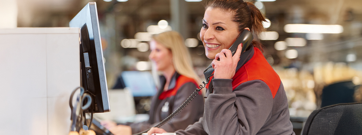 Eine Person in Selgros-Arbeitskleidung telefoniert an einem Arbeitsplatz, während im Hintergrund eine weitere Person und Computerbildschirme zu sehen sind.