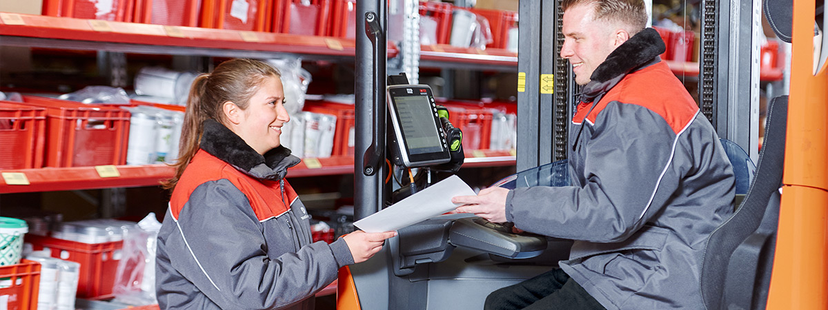 In einem Lager besprechen zwei Personen in Arbeitskleidung eine Liste, während eine Person auf einem Gabelstapler sitzt.