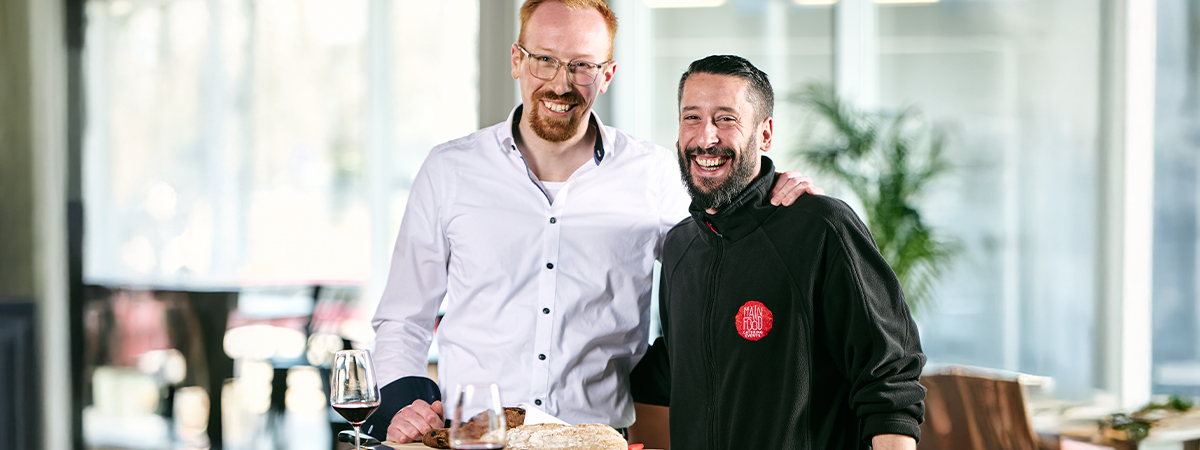 Zwei Personen stehen zusammen an einem Tisch, auf dem Wein, Brot und Speisen dekorativ arrangiert sind, in einer hellen und modernen Umgebung.