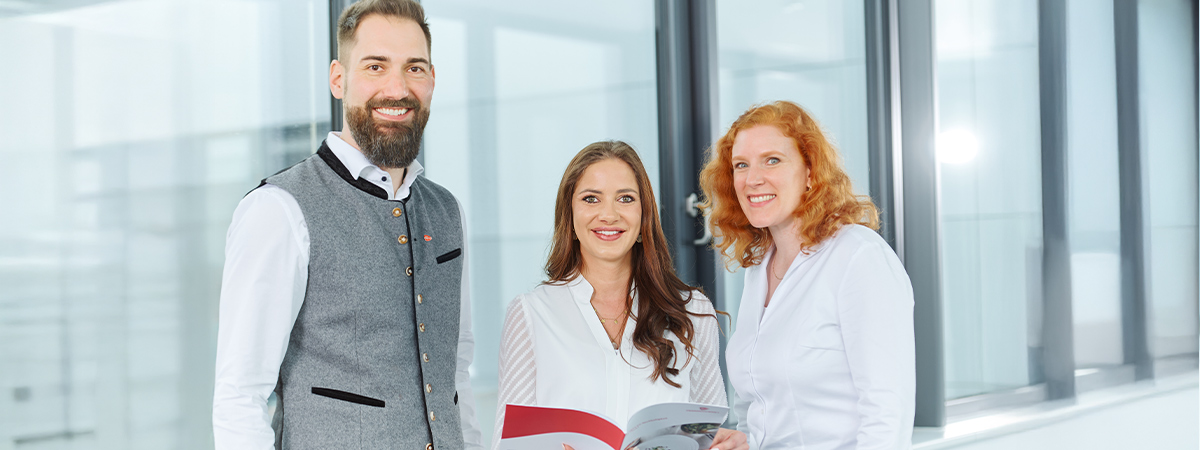 Drei Personen in Business-Kleidung stehen in einem modernen, hell beleuchteten Büro und betrachten gemeinsam ein Dokument.