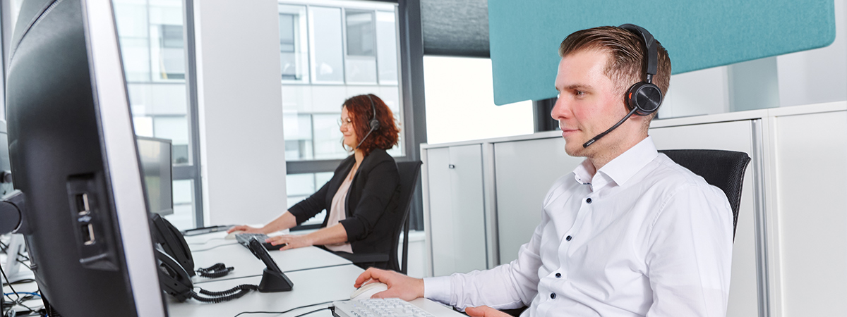 Zwei Personen arbeiten in einem modernen Büro an Computern, beide tragen Headsets.