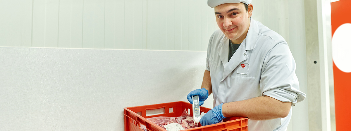 Eine Person in Arbeitskleidung kontrolliert die Temperatur des Fleisches in einem hygienischen Produktionsraum. Da Fleisch ist in einer rote Wannen zu sehen.