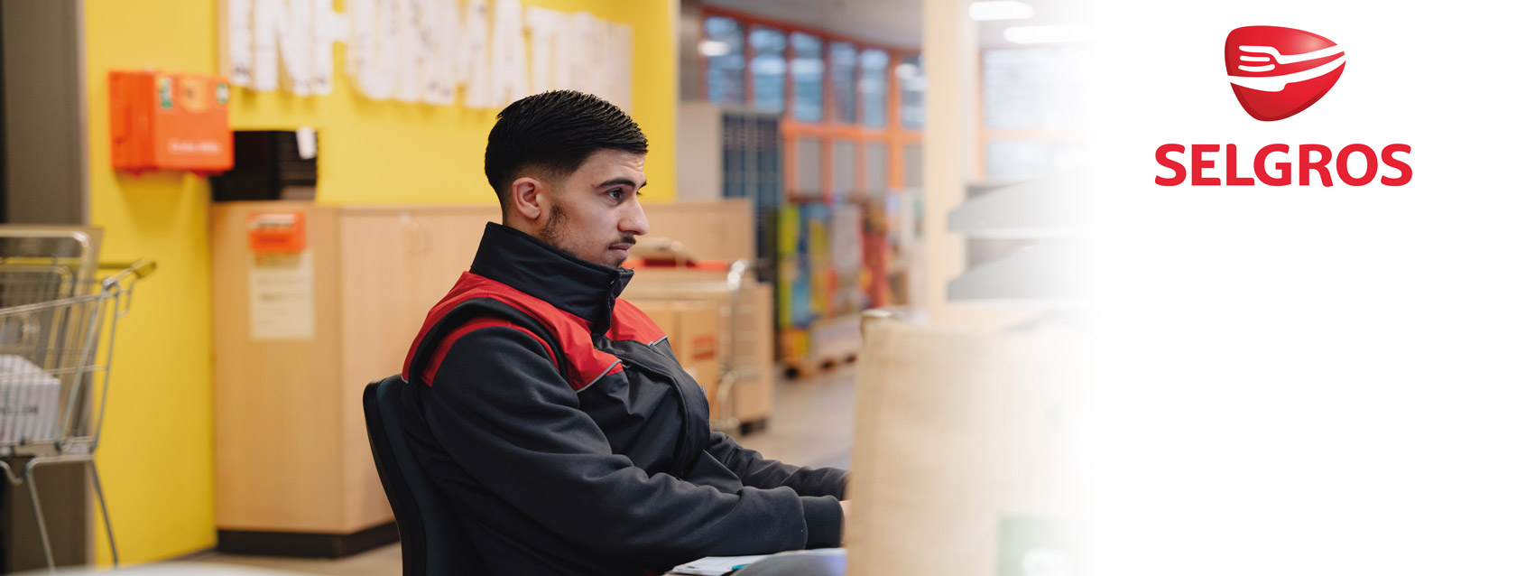Eine Person in Selgros-Arbeitskleidung sitzt an der Information einer Selgros-Filiale, während im Hintergrund Lagerbereiche und Aktenschränke zu sehen sind.