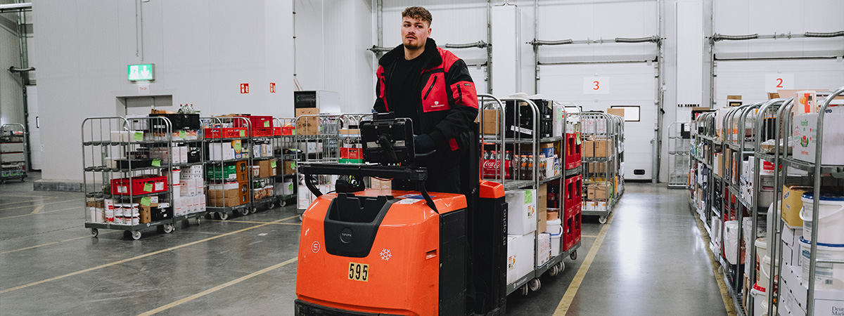 Eine Person arbeitet in einem Lager mit einem orangefarbenen Kommissionierfahrzeug, das mit Wagen voller Waren beladen ist.