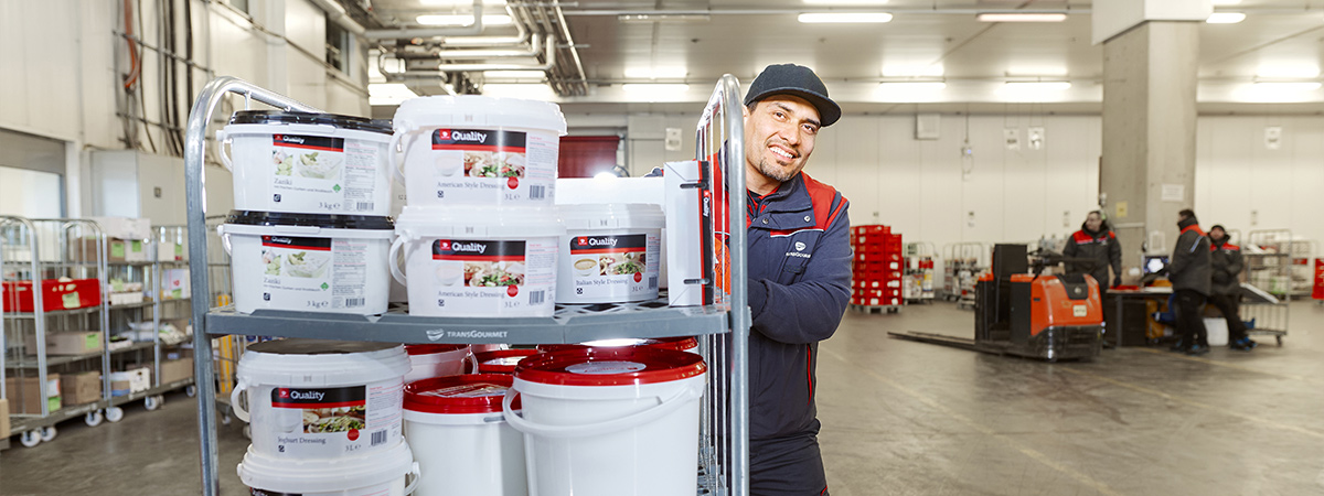 Eine Person in Arbeitskleidung steht in einem Lagerbereich neben einem Rollwagen, der mit großen Behältern und Eimern beladen ist. Im Hintergrund sind weitere Lageraktivitäten und orangefarbene Kommissionierfahrzeuge zu sehen.