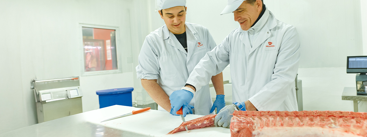 Zwei Personen stehen in weißer Arbeitskleidung in einer industriellen Umgebung. Eine Person schneidet ein Stück Fleisch während die andere Person dabei zuschaut.